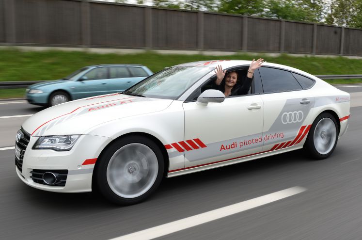 Eine Audi-Mitarbeiterin sitzt während einer Testfahrt auf dem Fahrersitz eines Audi, der über einen Testabschnitt auf der Autobahn A9 bei Ingolstadt fährt und winkt mit beiden Händen aus dem Fenster. 