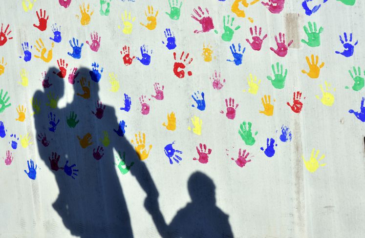 Ein Mann mit einem Kind auf dem Arm und einem an der Hand wirft am 08.04.2015 in München (Bayern) einen Schatten auf eine mit bunten Handabdrücken bemalte Wand einer Kindertagesstätte.