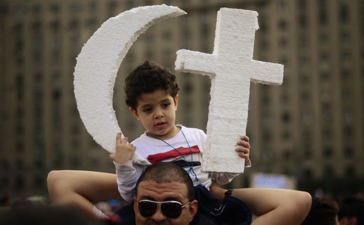 auf den Schultern eines Mannes sitzend hält ein kleiner Junge die beiden Glaubenssymbole "Kreuz" und "Halbmond" in der Hand 