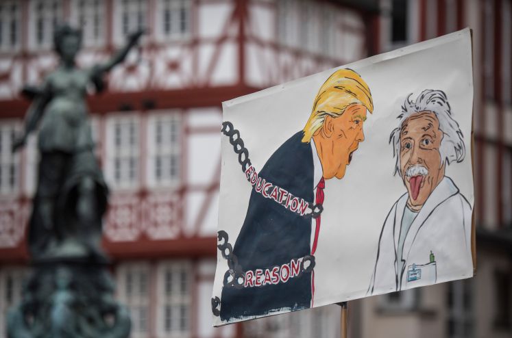 Ein Plakat wird hochgehalten. Darauf abgebildet Albert Einstein, die Zunge hinausstreckend. Neben ihm brüllt ihn Trump an, der in Ketten zurückgehalten wird. Auf den Ketten steht geschrieben: "Education" und "Reason"
