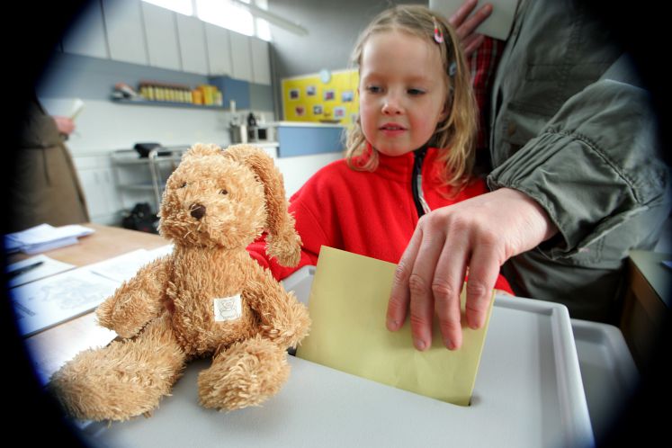Einwurf eines Stimmzettels in die Wahlurne. Daneben setzt ein Mädchen ihr Stofftier auf die Wahlurne.