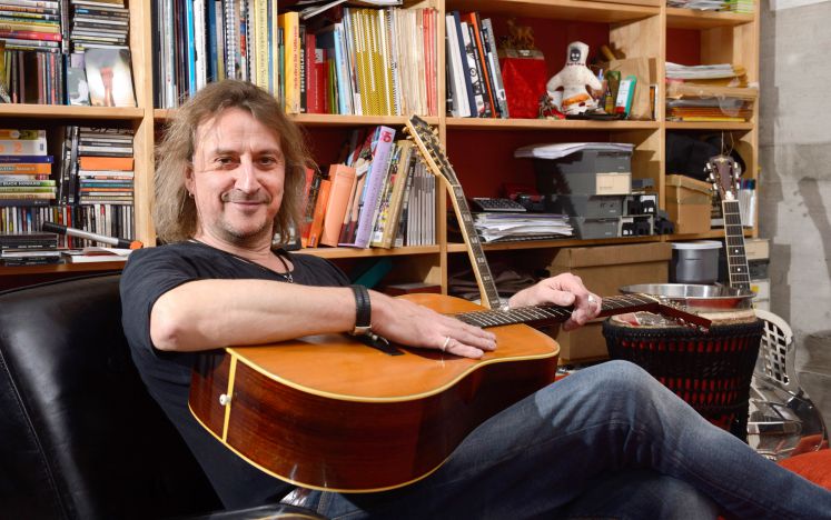 Dieter Thomas Kuhn sitzt mit seiner Gitarre auf einem Sessel und lächelt in die Kamera