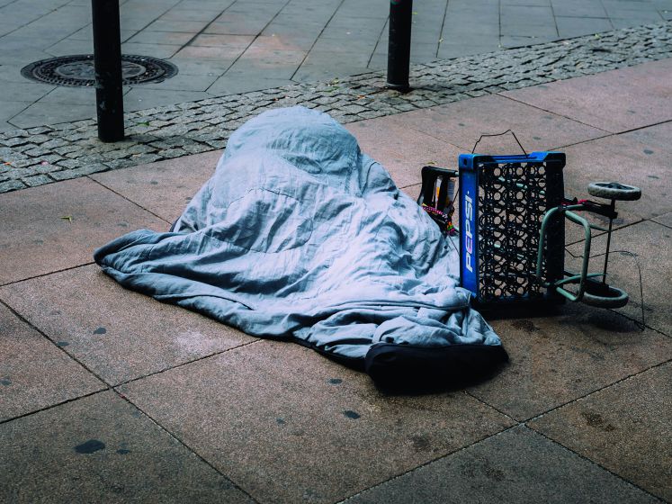 Illegaler Einwanderer auf der Straße, versteckt unter einer Decke, daneben ein umgekippter Einkaufswagen