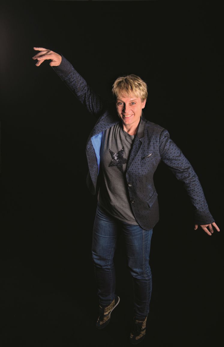 Jutta Kleinschmidt vor schwarzem Hintergrund in einer Pose wie eine auf dem Mond springende Astronautin