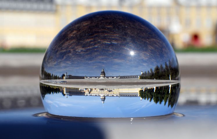 Baden-Württemberg, Karlsruhe: Das Karlsruher Schloss spiegelt sich in einer Glaskugel wider, die im Vordergrund auf der Wasserfläche einer Grünanlage liegt.