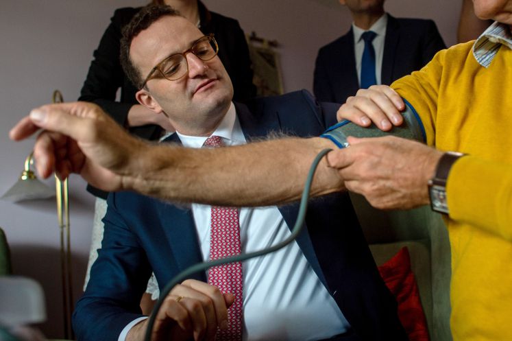 06.12.2019, Sachsen-Anhalt, Halle (Saale): Bundesgesundheitsminister Jens Spahn (CDU) hilft dem Probanden Norbert Neumann während einer Echt-Televisite mit einem Arzt beim anlegen einer manschette zum messen des Blutdrucks. In der Wohnung wird derzeit das Telemedizin-Projekt "Händel II" erprobt. Der Bundesgesundheitsminister besucht Sachsen-Anhalt auf Einladung des Ministerpräsidenten. 