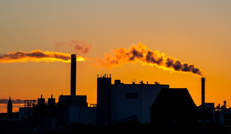 Rauchschwaden steigen aus einem Kamin auf. Die untergehende Sonne lässt die Wolken und den Rauch orange leuchten.