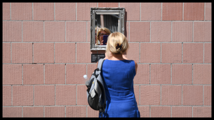 Eine blonde Frau in blauem Kleid steht vor Spiegel an roter Backsteinmauer