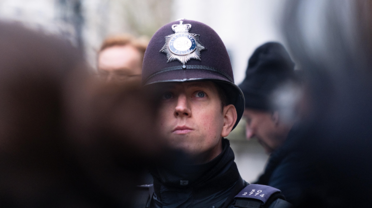 Britischer Polizist mit Helm in Uniform; unsicherer Vorschlag: weitere Personen im Hintergrund. Foto.