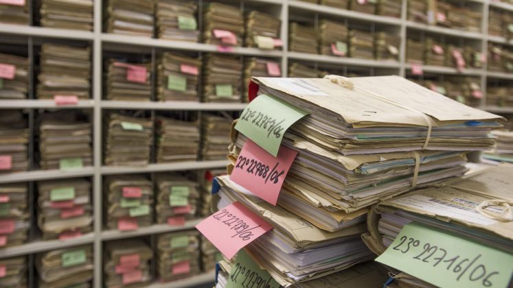 Stapel von Akten mit farbigen Zetteln in einem Archivraum, Regale voller weiterer Akten. Foto.