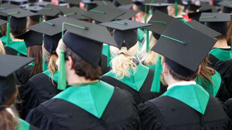 Foto: Viele Absolventen in schwarzen Roben und Hüten bei einer Abschlussfeier, Rückenansicht.