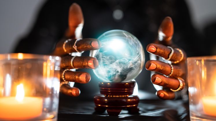 Foto: Zwei Kerzen und Hände mit Ringen um eine leuchtende Glaskugel auf dunklem Tisch.