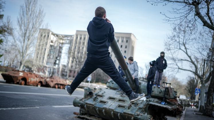 Foto: Junge Menschen klettern auf zerstörtem Panzer in urbaner Umgebung, Hochhaus im Hintergrund.