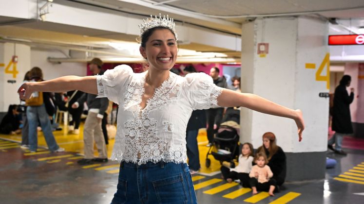 Frau mit Krone und Spitzenbluse tanzt lachend in Tiefgarage; weitere Menschen im Hintergrund. Foto.