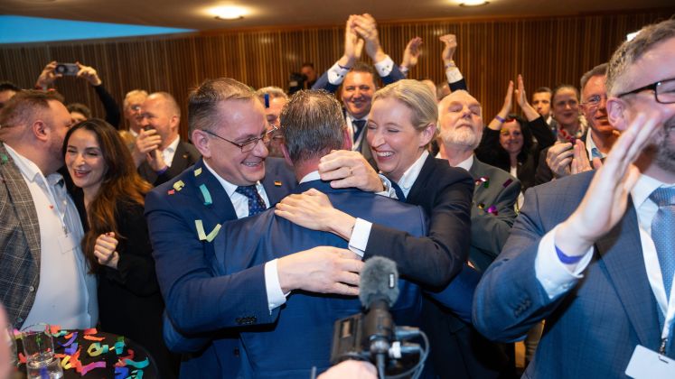 Gruppe feiert in festlichem Raum, mehrere Menschen umarmen sich und lachen, Farbfoto.