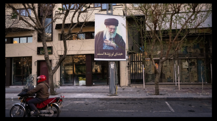 Foto: Motorradfahrer mit rotem Helm fährt an Plakat mit Porträt von Ali Khamenei vorbei.