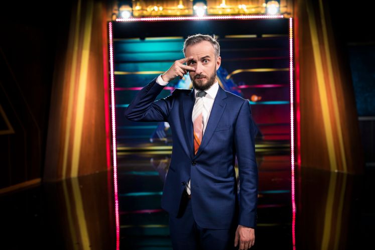 Jan Böhmermann im blauen Anzug vor bunter Lichtwand, posiert mit Hand vor dem Gesicht, Foto.