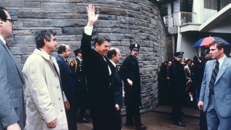 Ronald Reagan winkt im Anzug, umgeben von Männern in Anzügen und Polizei; Foto, draußen.