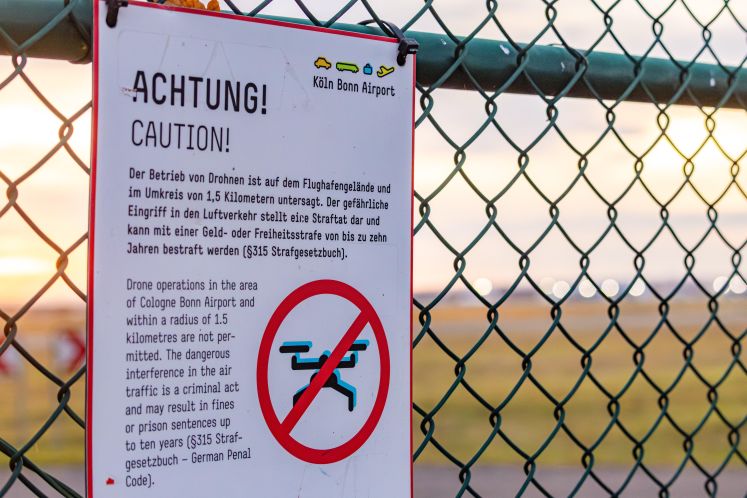 Foto eines Warnschilds an einem Zaun, verbietet Drohnen, unscharfer Flughafen im Hintergrund.