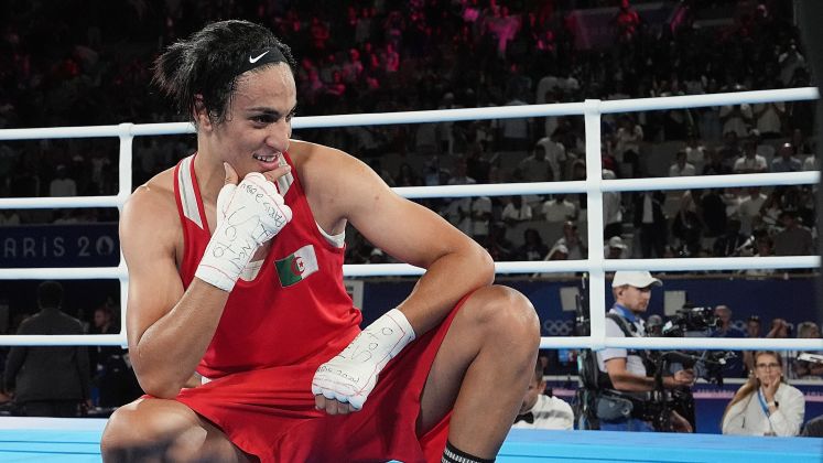 Boxerin im roten Trikot kniet lächelnd im Boxring; Publikum im Hintergrund, Foto.