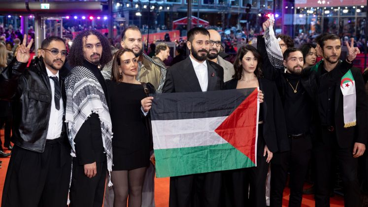 Mehrere Menschen auf rotem Teppich halten eine palästinensische Flagge, einige zeigen das Peace-Zeichen. Foto.