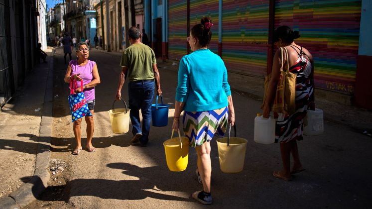 Vier Menschen tragen Wasserkanister auf einer sonnigen Straße, bunte Wand im Hintergrund, Foto.