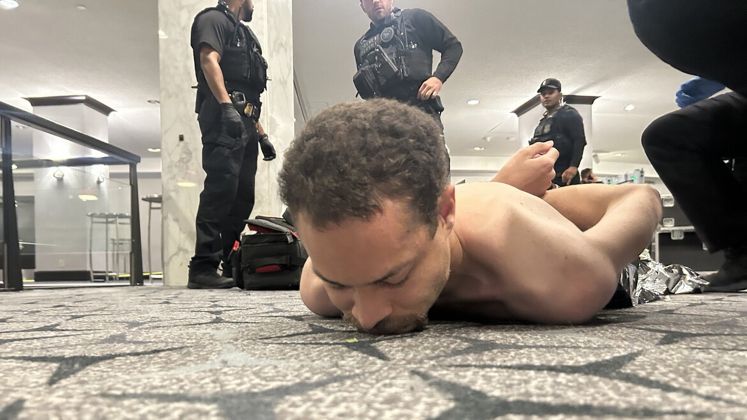 Oberkörperfreier Mann liegt gefesselt am Boden, mehrere Polizisten stehen herum, Innenraum, Foto.