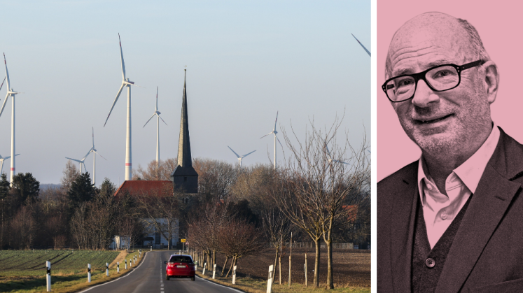 Foto: Landstraße mit rotem Auto, Windräder und Kirchturm im Hintergrund, keine Personen erkennbar.
