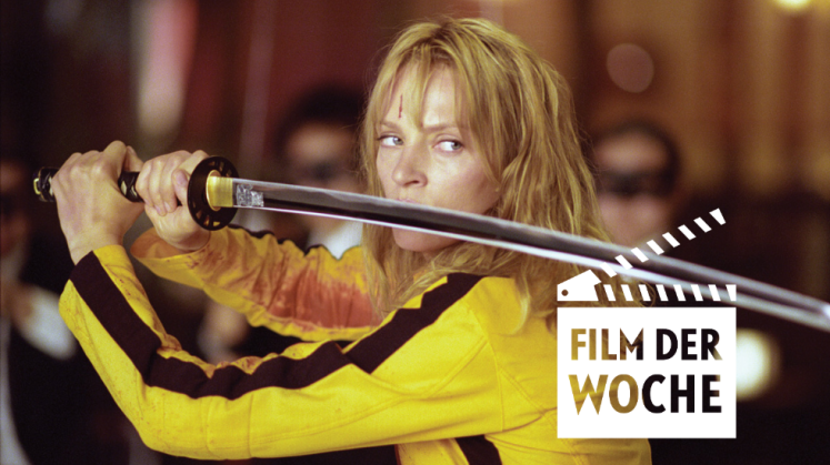 Foto: Uma Thurman im gelben Kampfanzug hält ein Katana, angespannt, unscharfer Hintergrund.