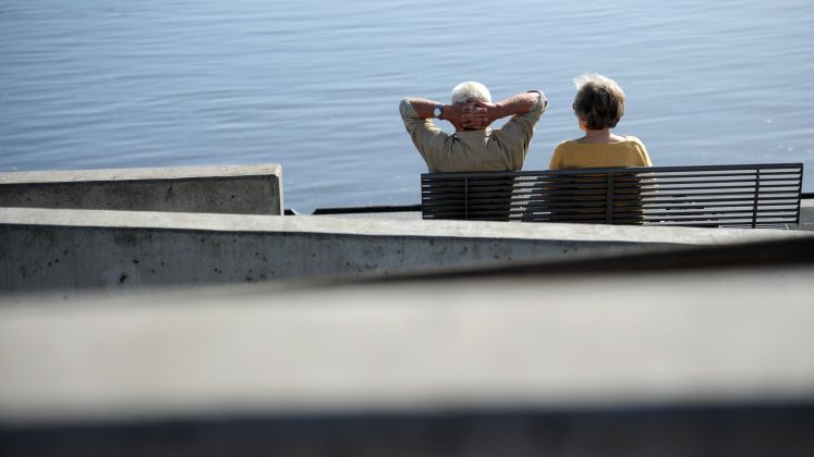Zwei ältere Personen sitzen entspannt auf einer Bank am Wasser, Foto.