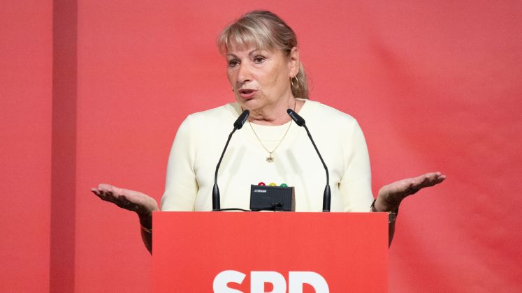 Frau am SPD-Rednerpult gestikuliert im cremefarbenen Blazer vor rotem Hintergrund, Foto.