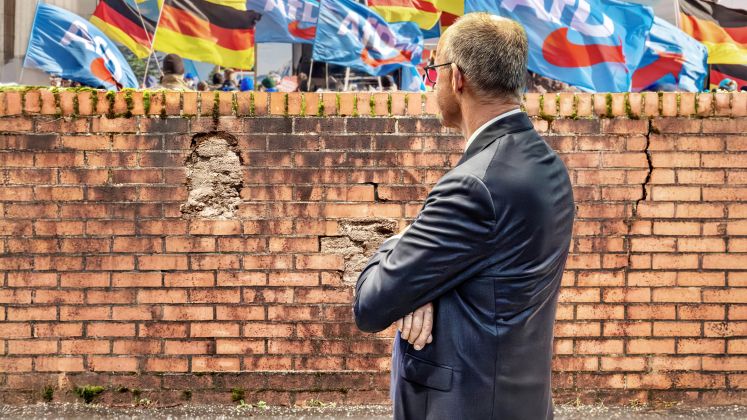 Merz im Anzug steht vor Backsteinmauer, im Hintergrund Deutschland- und AfD-Flaggen