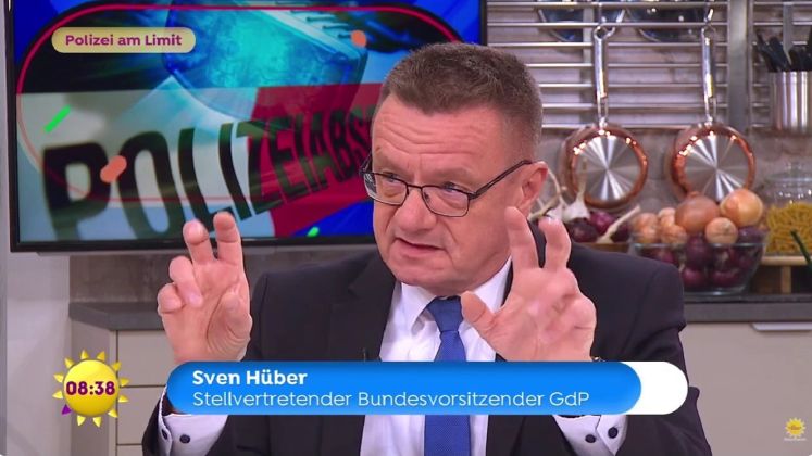 Foto: Sven Hüber im Studio, gestikulierend, im Hintergrund Polizeikelle und Dekoration.