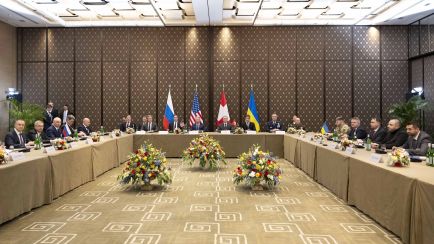 Große Gruppe von Politikern an einem runden Tisch mit Blumen, russischer und ukrainischer Flagge, Foto.
