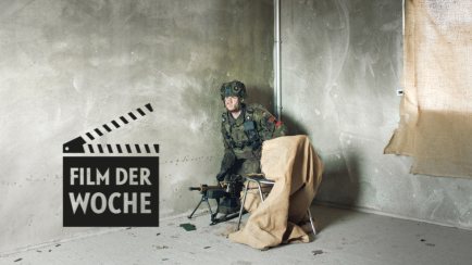 Soldat in Uniform sitzt mit Gewehr an der Wand in kargem, grauem Raum