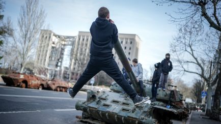 Foto: Junge Menschen klettern auf zerstörtem Panzer in urbaner Umgebung, Hochhaus im Hintergrund.