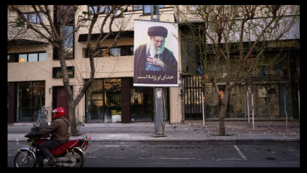 Foto: Motorradfahrer mit rotem Helm fährt an Plakat mit Porträt von Ali Khamenei vorbei.