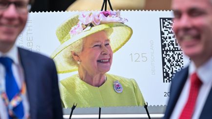 Briefmarke mit Queen Elizabeth II. im gelben Kleid und Hut, zwei unscharfe Männer im Vordergrund, Foto.