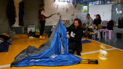Eine Person baut ein Zelt in einem Innenraum ab, drei weitere Personen sitzen im Hintergrund. Foto.