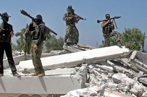 Vier bewaffnete, maskierte Männer stehen auf Trümmern; sonnige Außenaufnahme, Foto.