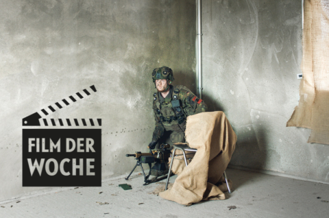 Soldat in Uniform sitzt mit Gewehr an der Wand in kargem, grauem Raum