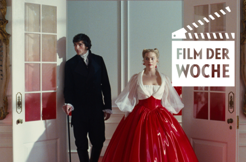 Mann im schwarzen Anzug und Frau im roten Kleid betreten prunkvollen Raum