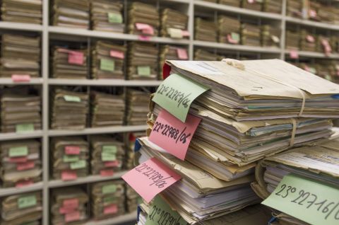 Stapel von Akten mit farbigen Zetteln in einem Archivraum, Regale voller weiterer Akten. Foto.