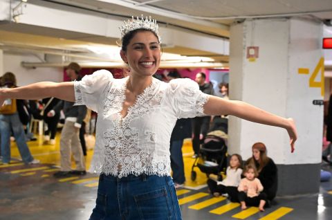 Frau mit Krone und Spitzenbluse tanzt lachend in Tiefgarage; weitere Menschen im Hintergrund. Foto.