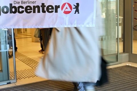 Foto: Person mit hellem Mantel betritt ein Gebäude, darüber Banner mit „jobcenter“-Logo.