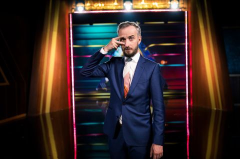 Jan Böhmermann im blauen Anzug vor bunter Lichtwand, posiert mit Hand vor dem Gesicht, Foto.