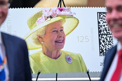 Briefmarke mit Queen Elizabeth II. im gelben Kleid und Hut, zwei unscharfe Männer im Vordergrund, Foto.