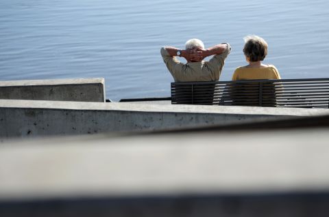Zwei ältere Personen sitzen entspannt auf einer Bank am Wasser, Foto.