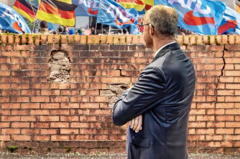 Merz im Anzug steht vor Backsteinmauer, im Hintergrund Deutschland- und AfD-Flaggen