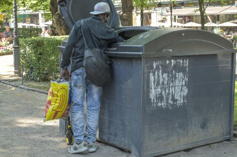 Mann mit Basecap sucht in einem großen Müllcontainer in einem Park, Einkaufstüte in der Hand. Foto.